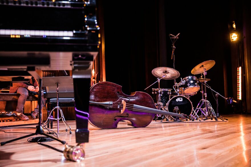 A jazz stage set for performance featuring a double bass lying on its side beside a drum kit, with part of a grand piano visible in the foreground. Warm stage lighting reflects across the polished wooden floor in a modern concert venue.  Photo credit: Photo by Dolo Iglesias / Unsplash
