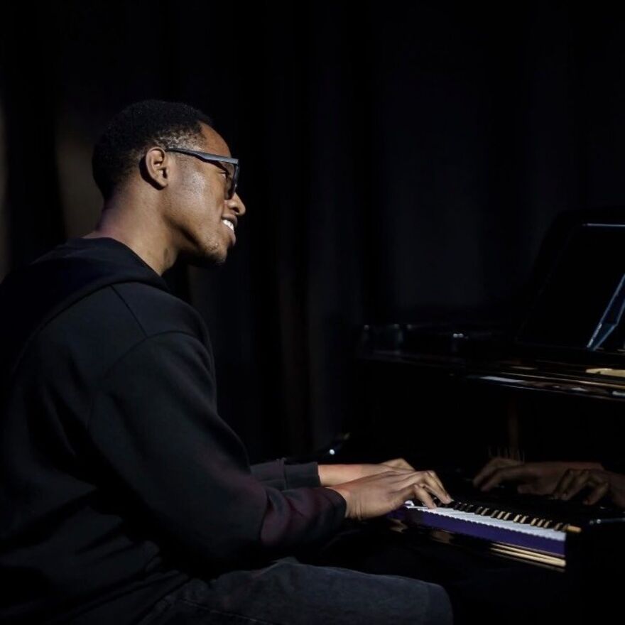 young male sitted at piano wearing glasses and smiling in black environment