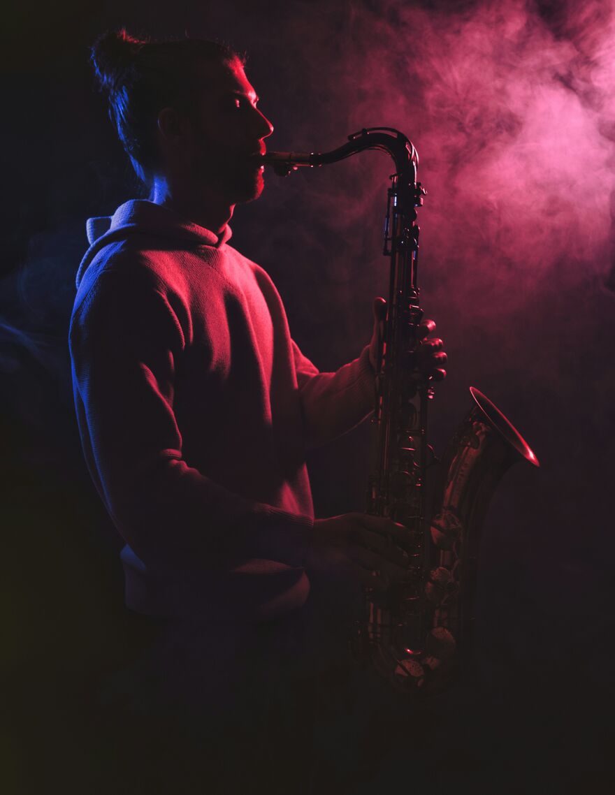 Man with pony tail and sweat shirt plaving saxophone in dark room with pink smoke